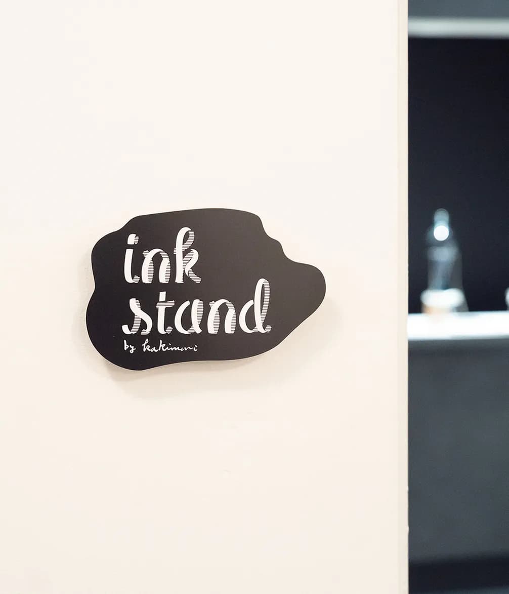 A black, cloud-shaped sign with "ink stand by kakimori" in white script hangs on a beige wall, next to a dark, blurry room.