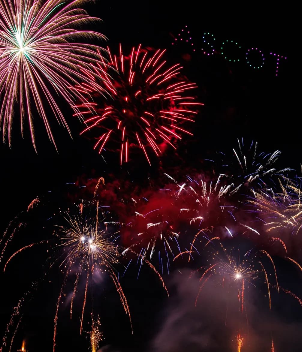 A vibrant fireworks display with bursts of red, green, and gold set against a dark night sky, spelling "ASCOT" in colorful lights.