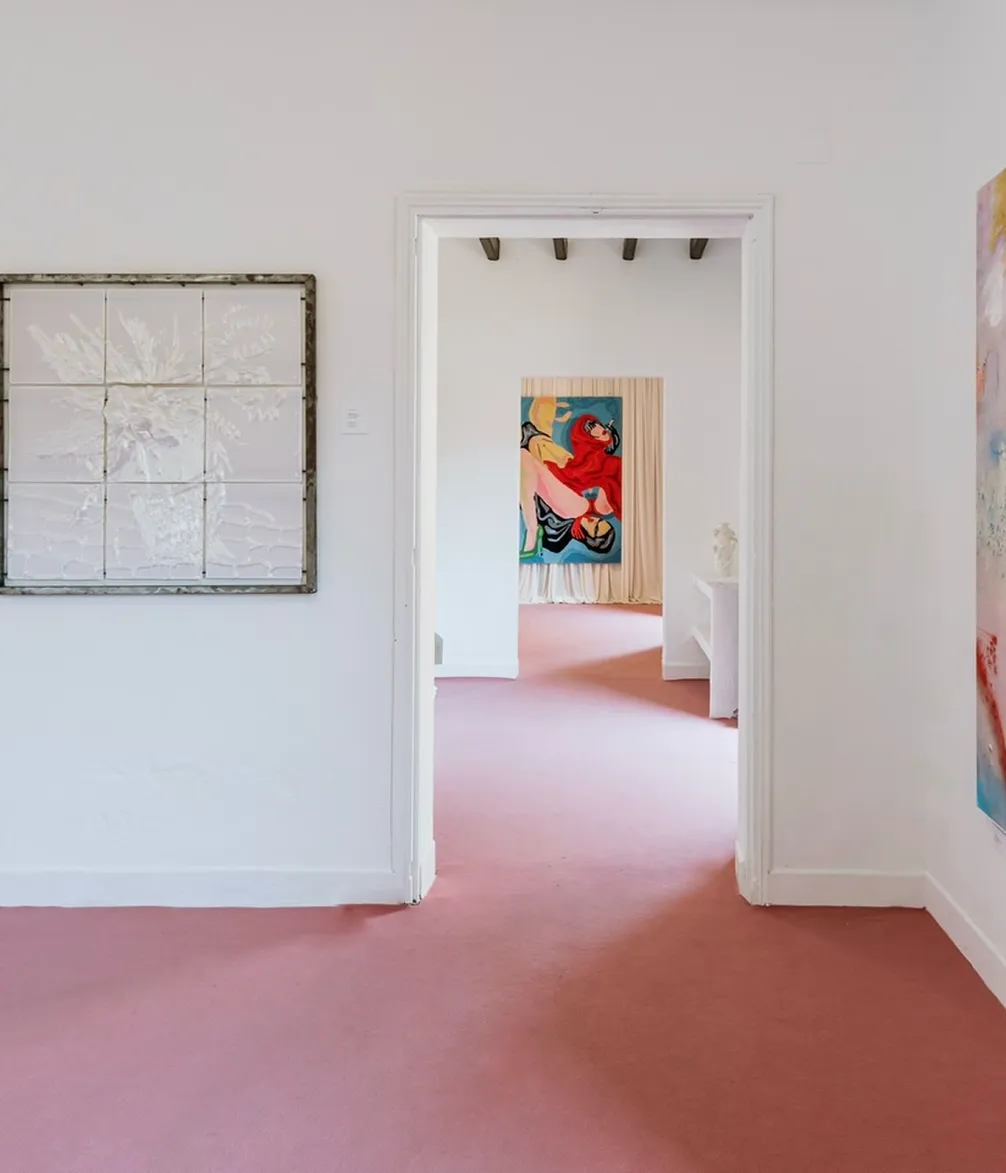 A bright room with pink carpet and white walls, featuring abstract artwork on the walls. A doorway reveals another room with a colorful painting.