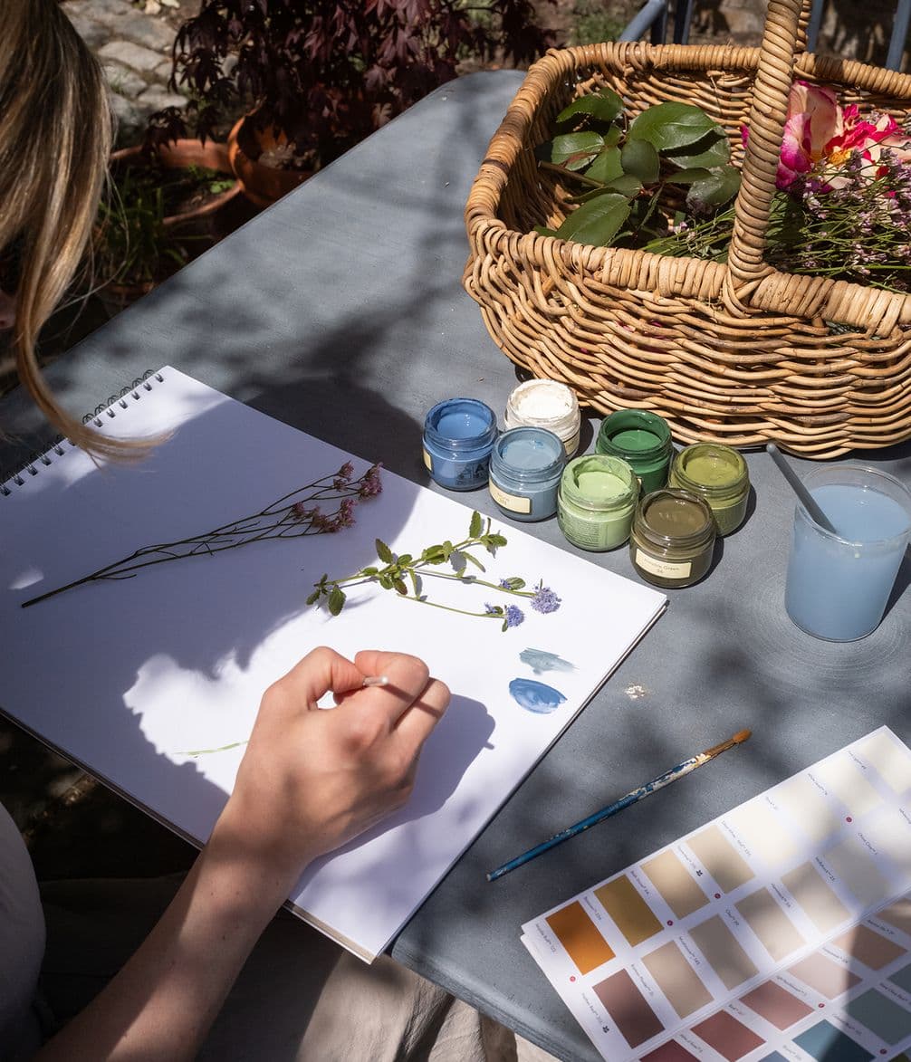 Person sketching flowers on paper outdoors, surrounded by paints, a brush, a color palette, and a wicker basket with pink and green plants.