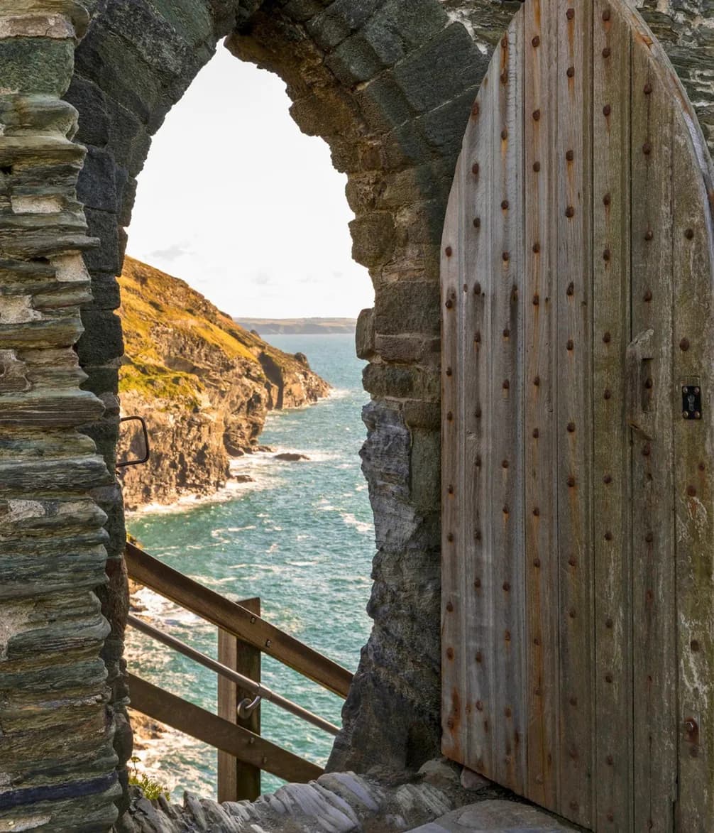 Stone archway with a wooden door overlooking a coastal cliff and the ocean beneath a clear sky.