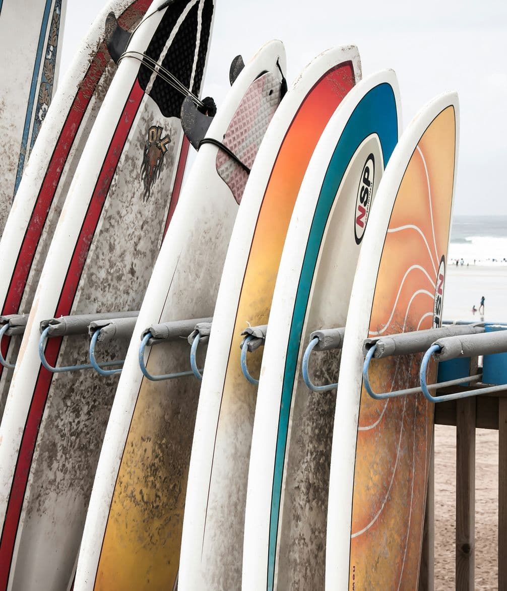 Surfboards of various colors and designs are lined up on a rack by the beach, with the ocean visible in the background.