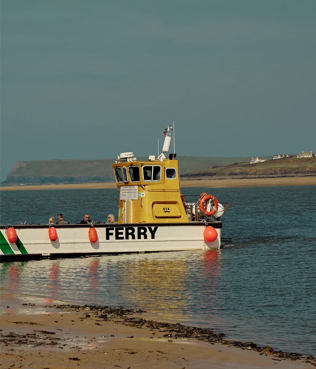 A yellow ferry with passengers onboard navigates near the shore against a backdrop of distant hills and houses.