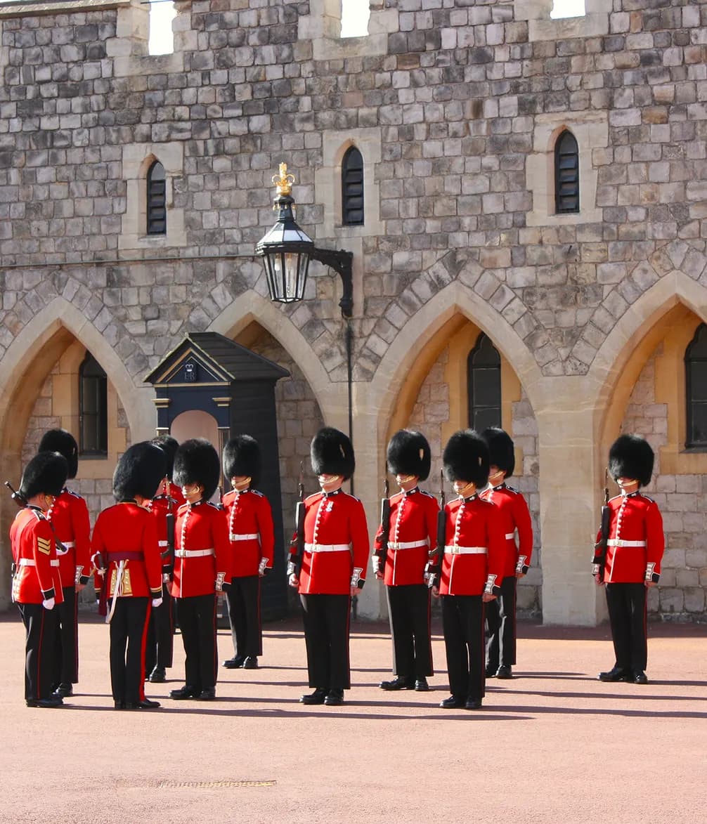 Guards in red uniforms and bearskin hats stand in formation outside a historic stone building.