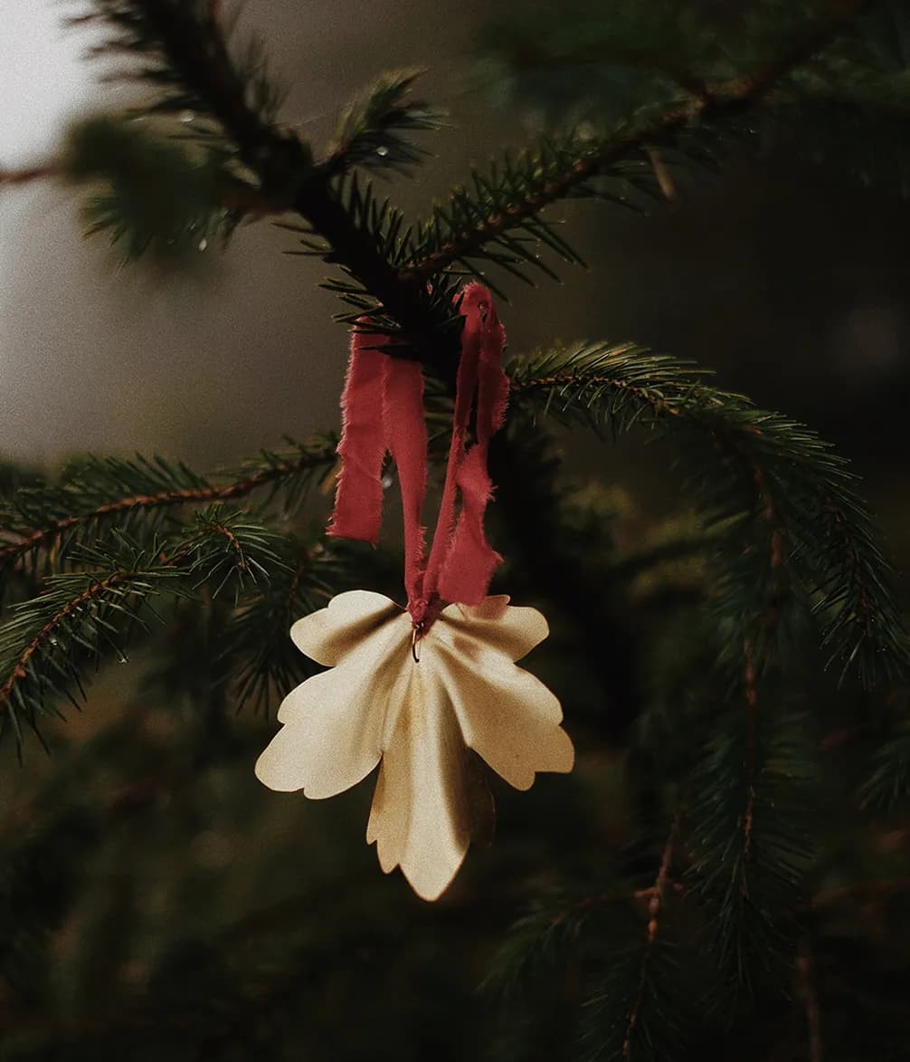 A paper flower ornament with red ribbon hangs on a green pine branch, against a blurred natural background.