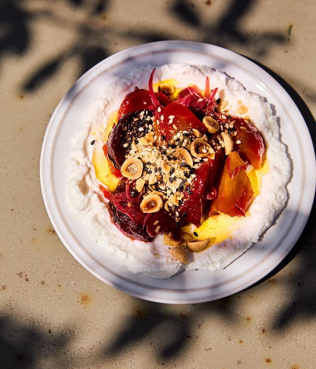 Plate of whipped cream topped with sliced roasted beets, toasted nuts, and seeds, garnished with vibrant red and yellow accents, on a light surface.