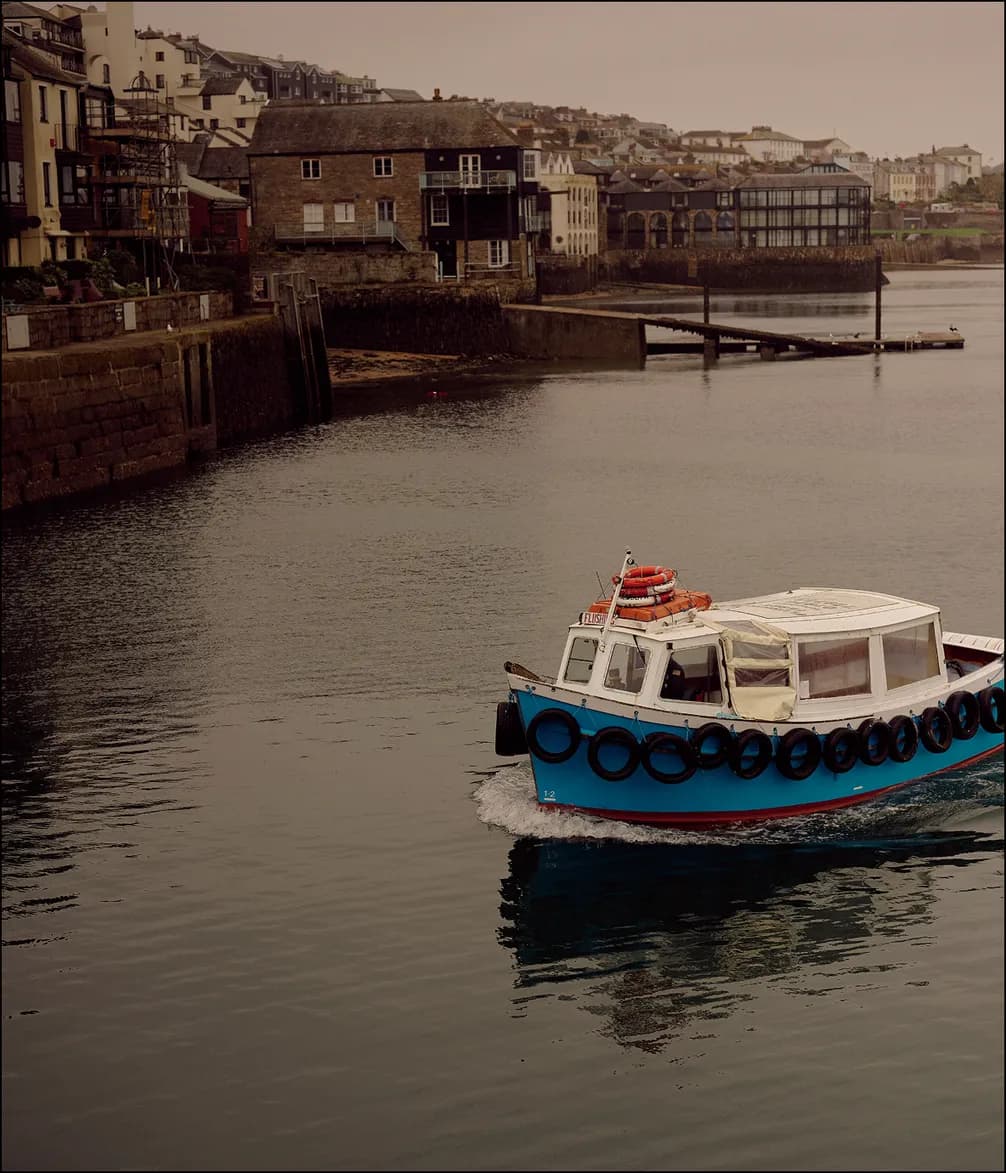 A blue and white boat with lifebuoys sails in a calm harbor near a row of seaside buildings under an overcast sky.