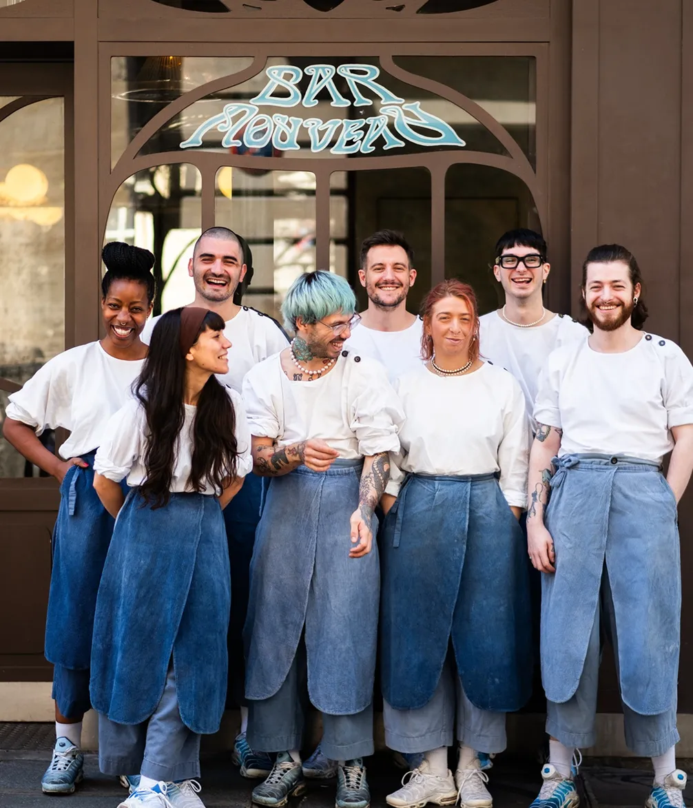 Group of eight people wearing matching blue denim aprons standing outside a bar, smiling and posing for the photo.