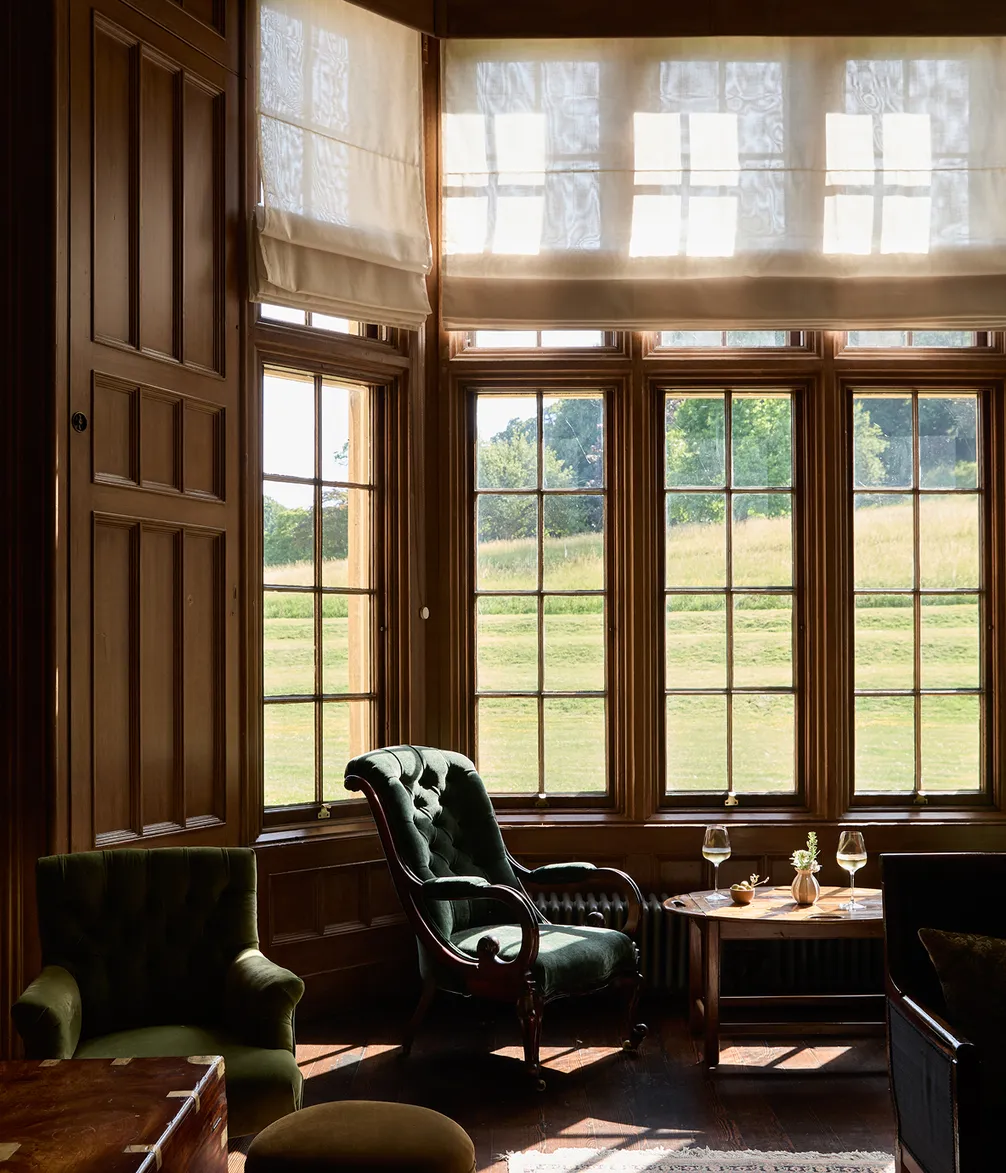Cozy room with large windows, warm sunlight spilling over a wooden floor, two green upholstered chairs, and a small table with wine glasses.