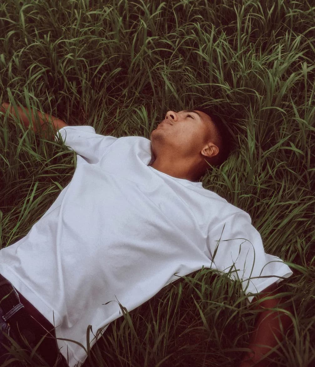 A person in a white t-shirt lies on their back in tall, lush green grass, looking relaxed and contemplative.