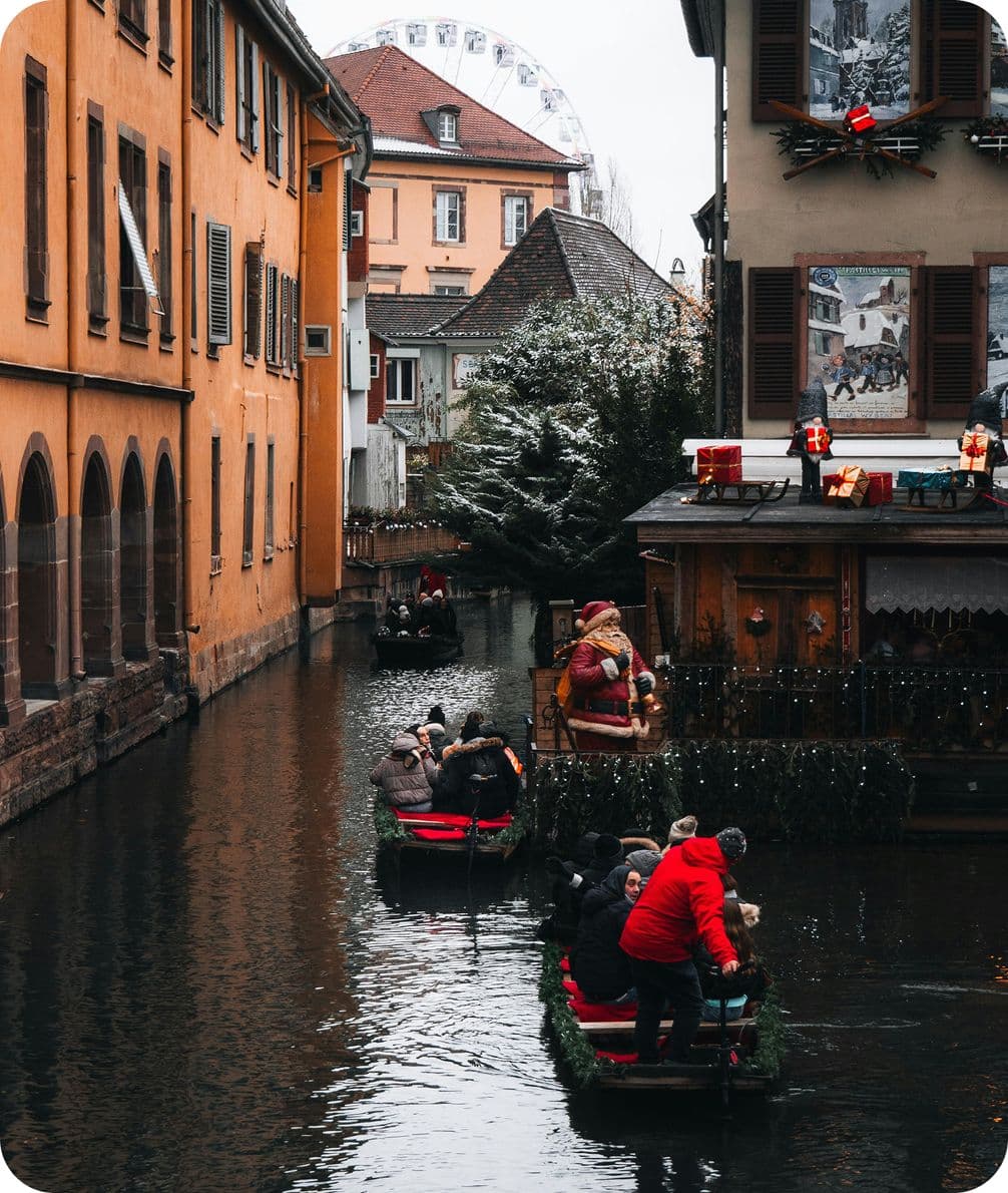 People in boats navigate a narrow canal flanked by festive buildings, with a Ferris wheel visible in the background.