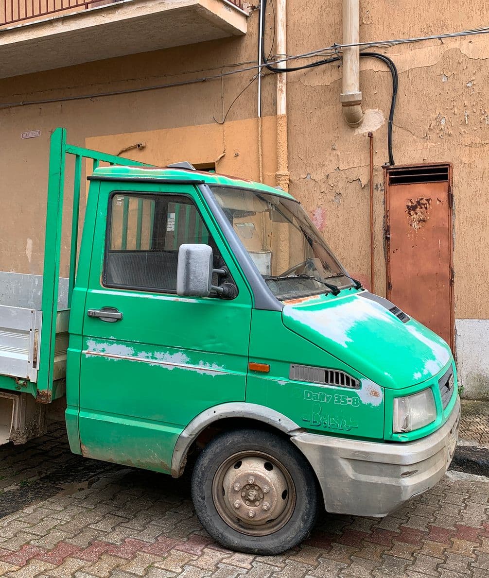 Green, weathered truck parked on a narrow street beside a tall, textured orange building with visible pipes and a rusty door.