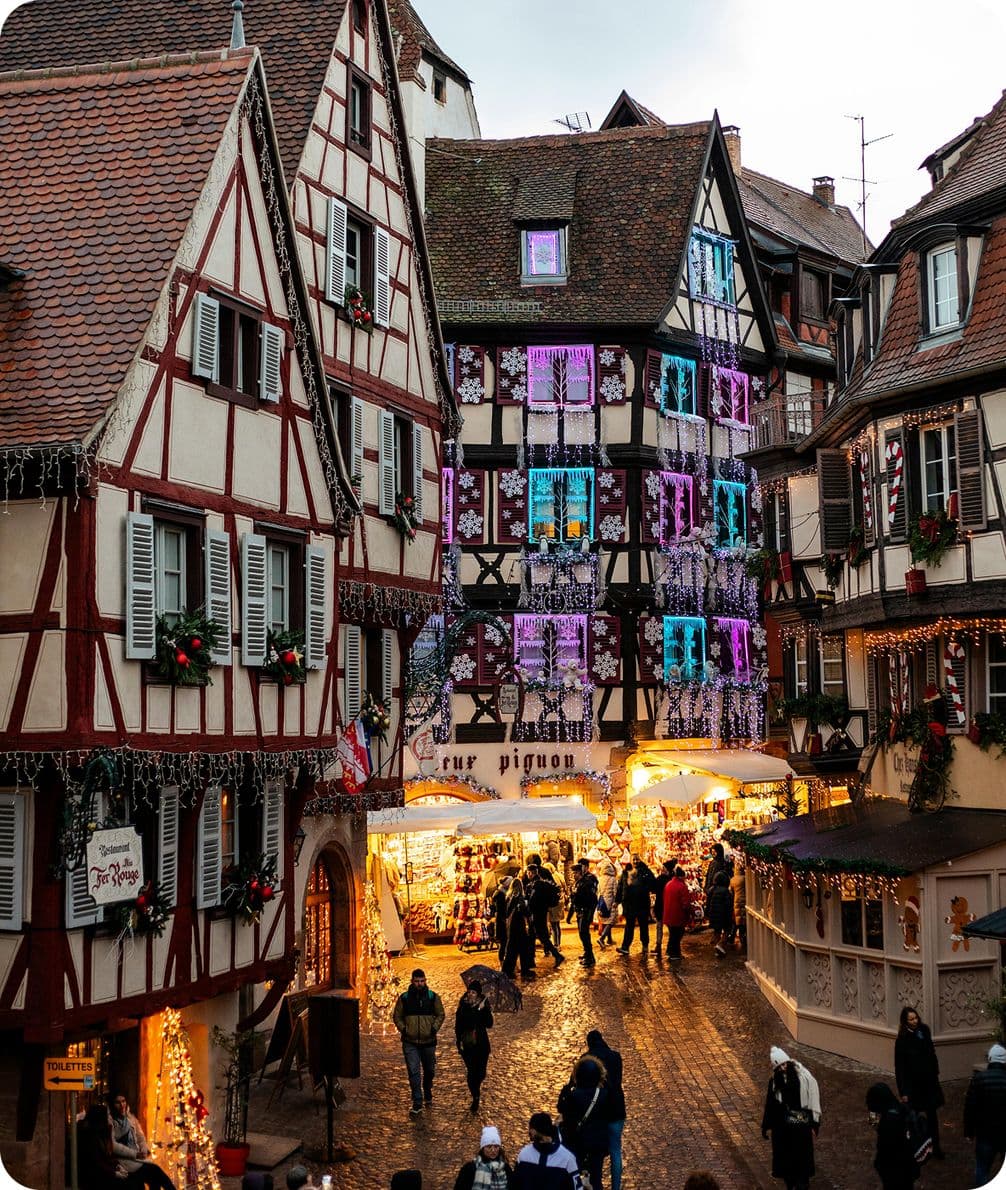 Charming European street with half-timbered buildings adorned with festive lights, bustling with people and a cozy shop display.