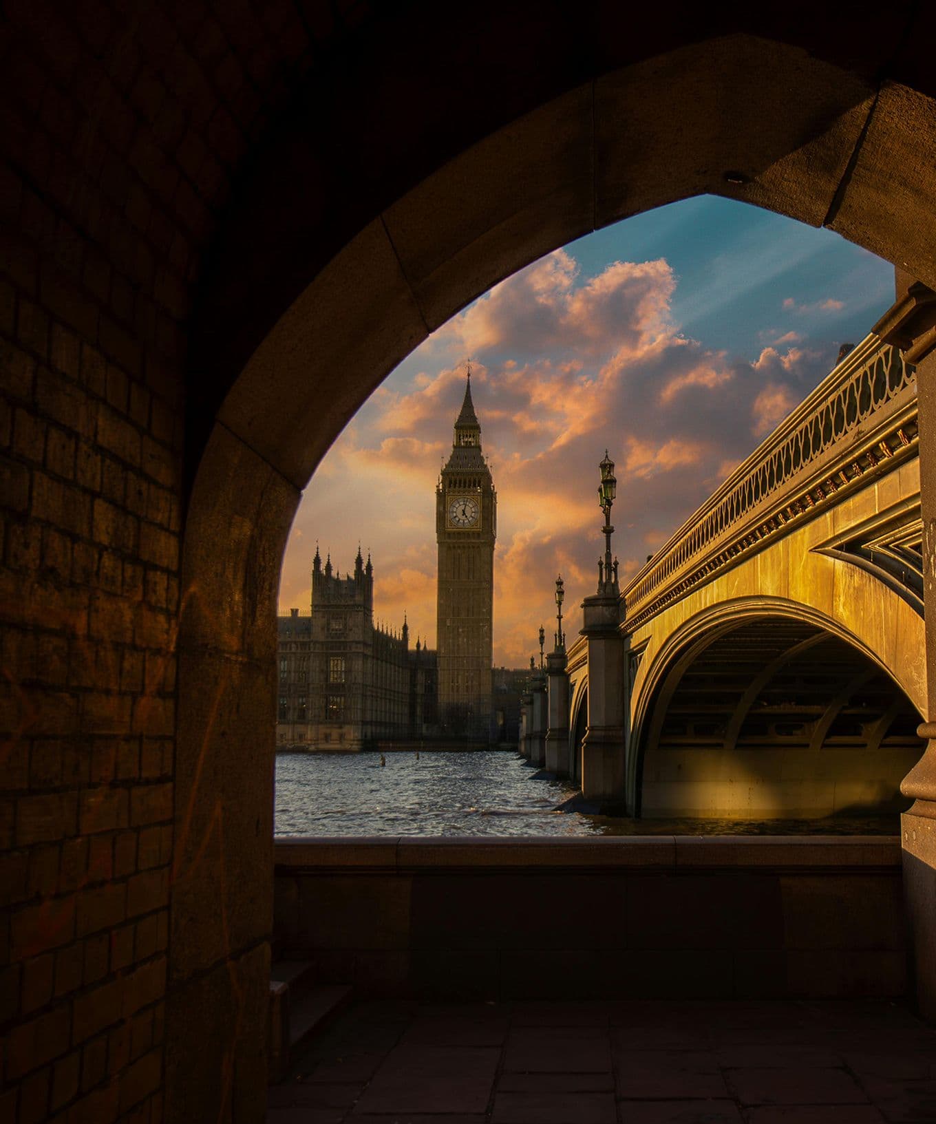 Arched view of Big Ben and Westminster Bridge over the Thames at sunset, with golden clouds and reflective water.