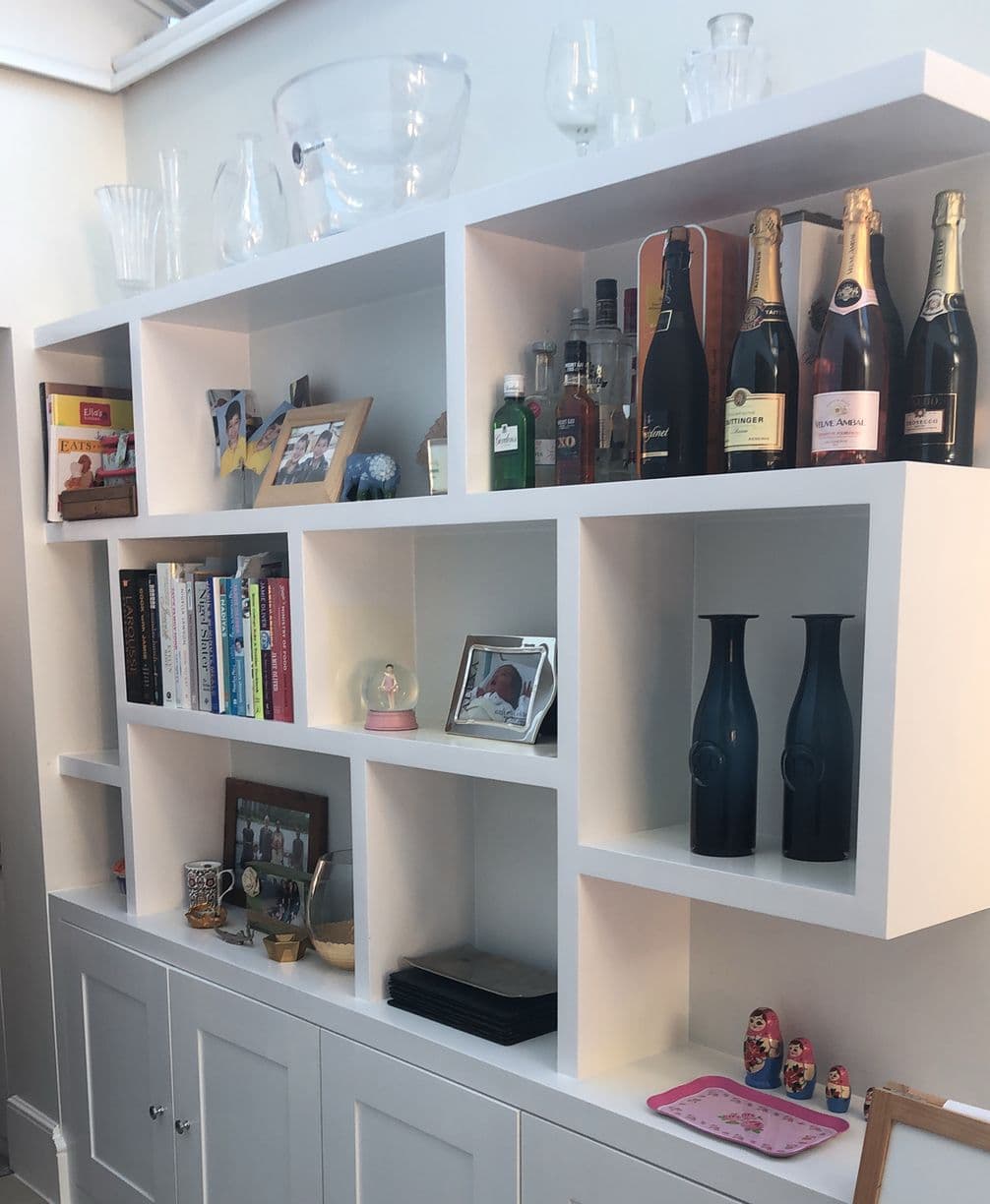 White modular shelving unit filled with books, framed photos, liquor and champagne bottles, glassware, vases and small decorative items.