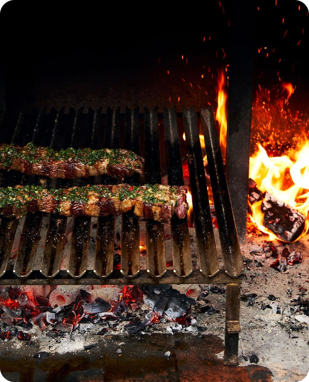 Two herb-seasoned steaks searing on a metal grill above glowing coals, with flames leaping to the right.