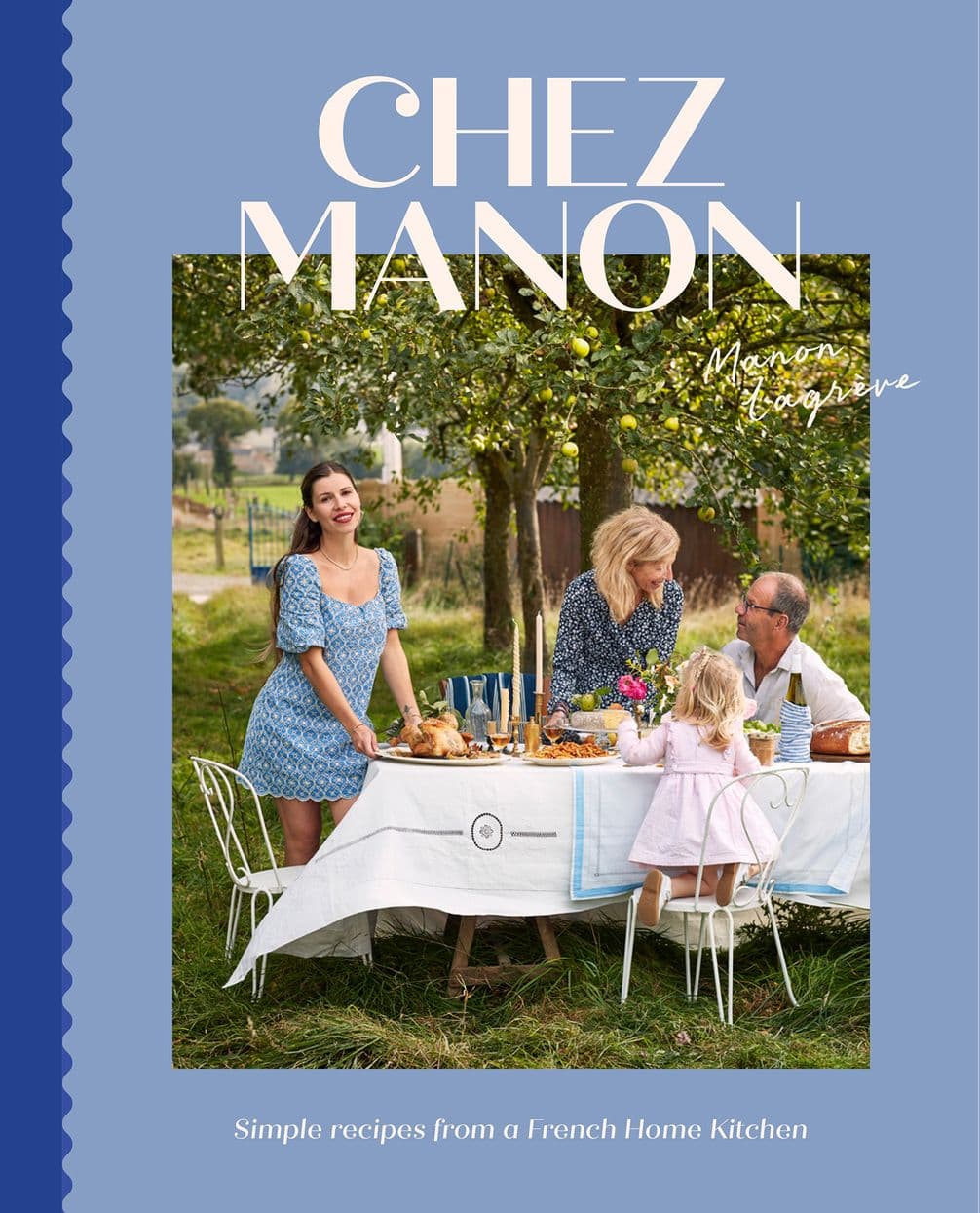 A family enjoys an outdoor meal under a tree. A woman in a blue dress stands smiling, while two adults and a child sit at a table with food.