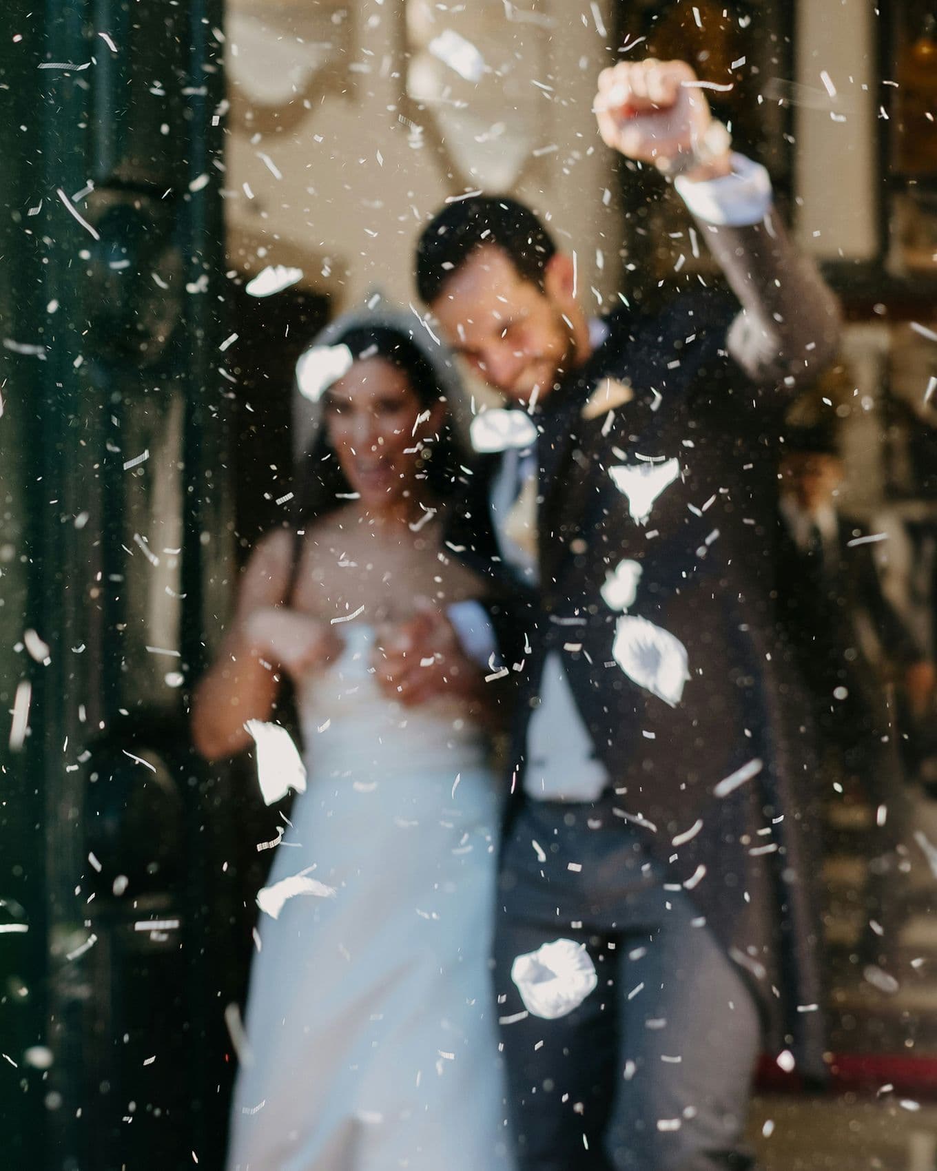 A joyous couple celebrates outside, surrounded by falling confetti. The bride holds her dress, and the groom raises his fist in excitement.