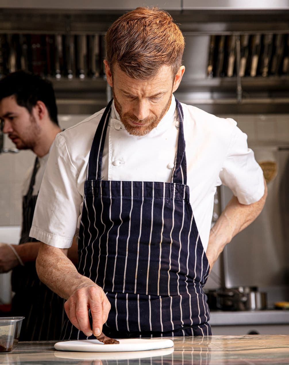 Chef in a striped apron carefully plates food in a professional kitchen, with another person working in the background.