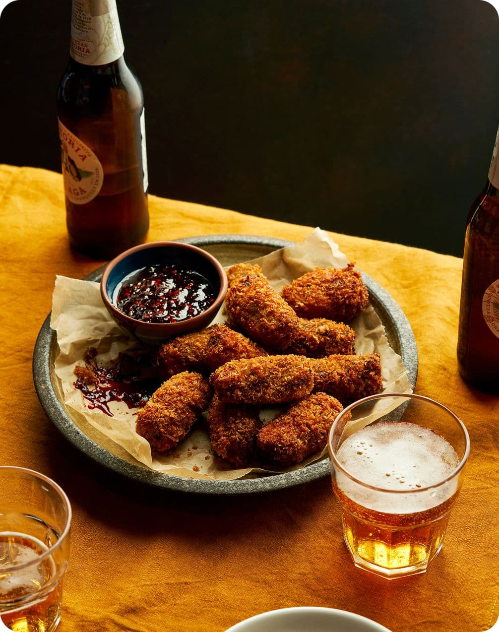 Plate of golden breaded croquettes with dark dipping sauce, beer bottles and glasses on a mustard-yellow tablecloth.