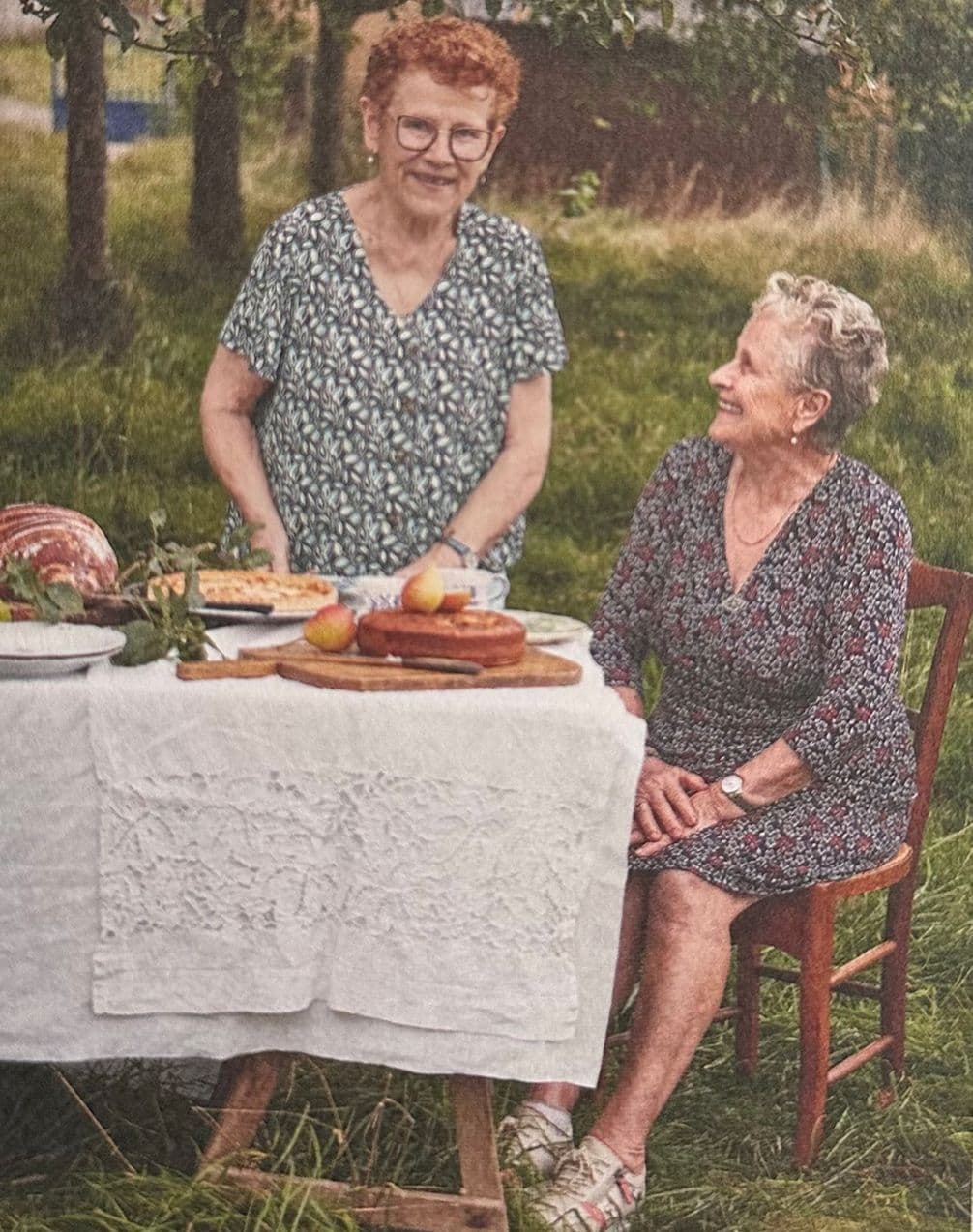Two elderly women in floral dresses enjoy an outdoor picnic with bread and fruits on a table, surrounded by greenery.