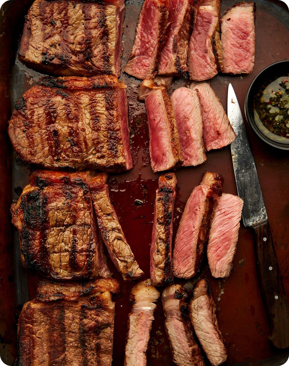 Assorted grilled steaks, sliced medium-rare on a dark tray with a carving knife and small bowl of herb sauce.