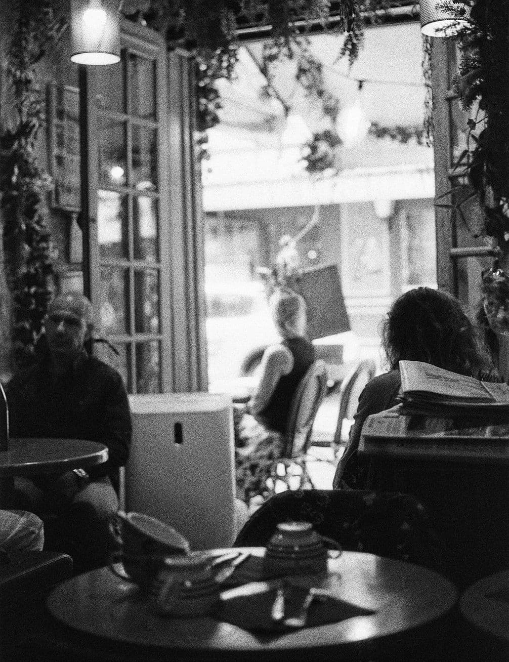 Black-and-white cafe interior with patrons at tables near an open window, teapot and cups on a foreground table, hanging plants and soft light.