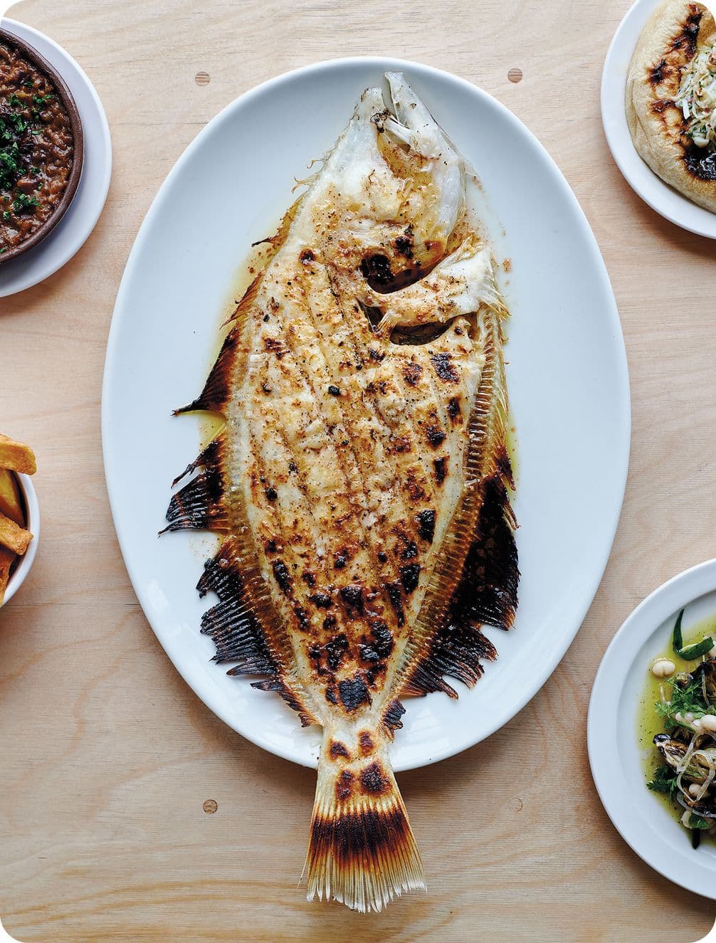 Grilled whole fish on a white oval plate, surrounded by side dishes on a wooden table.