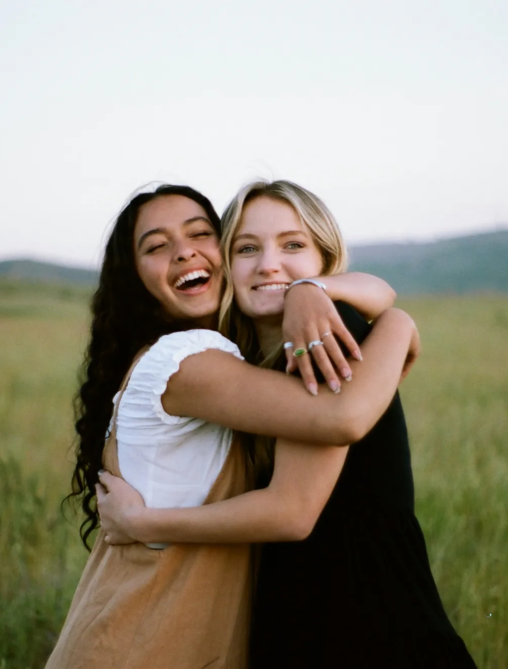 Two women smiling and hugging in a grassy field under a clear sky.