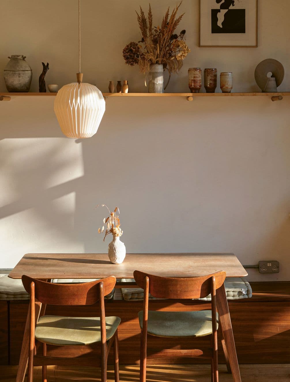 A cozy dining area with a wooden table and two chairs. A shelf with dried flowers and ceramics is above, and a pendant lamp hangs overhead.