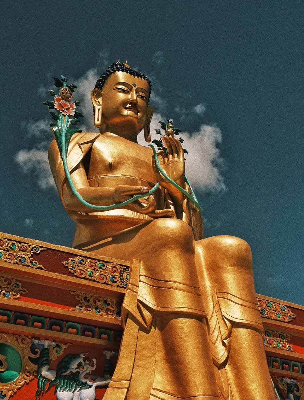 Golden statue of a seated figure holding flowers against a blue sky with clouds, adorned with ornate details and vibrant colors.