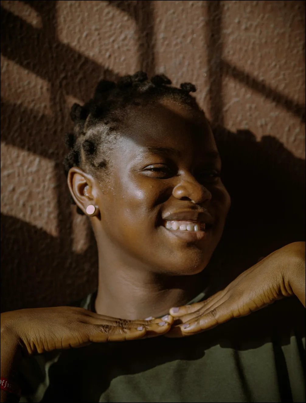 Smiling person with short braided hair and earrings, posing with hands under chin, against a textured wall with shadow patterns.