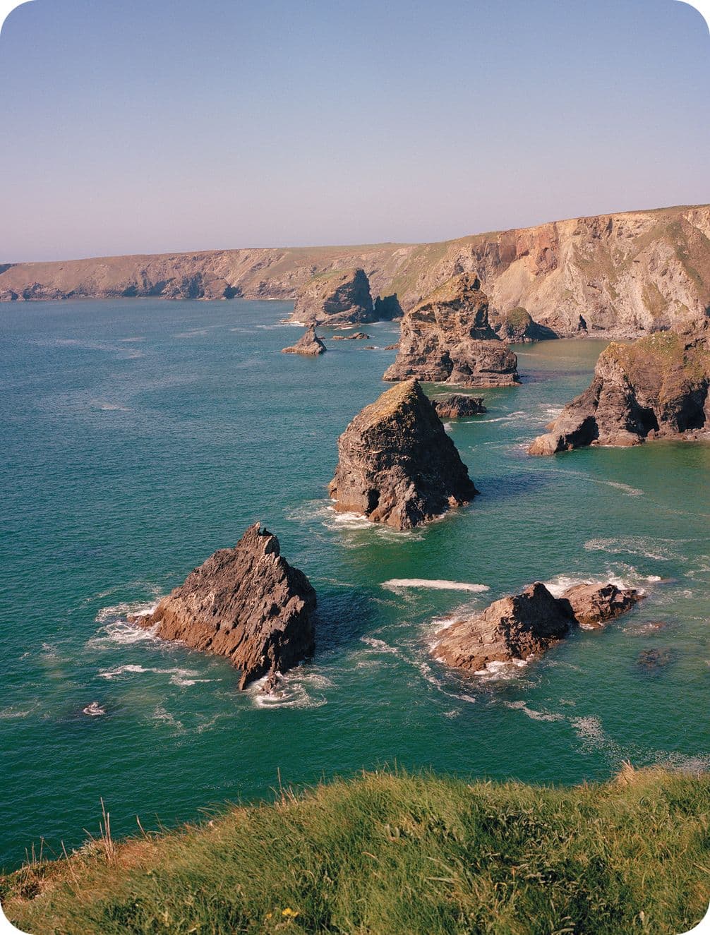 Coastal landscape with rugged cliffs and rocky sea stacks jutting out of the turquoise ocean under a clear blue sky.
