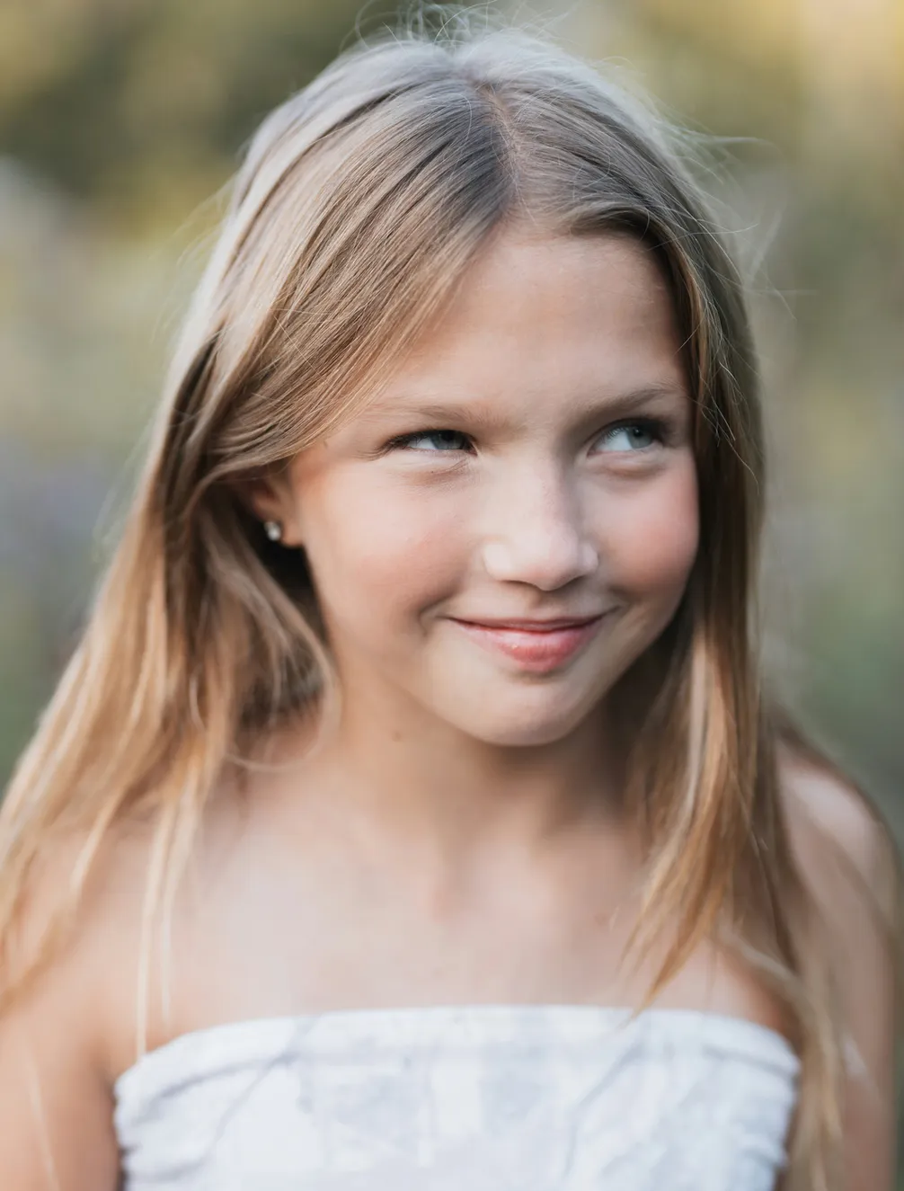 A young girl with long blonde hair and a mischievous smile stands outdoors, wearing a strapless top, with a blurred natural background.