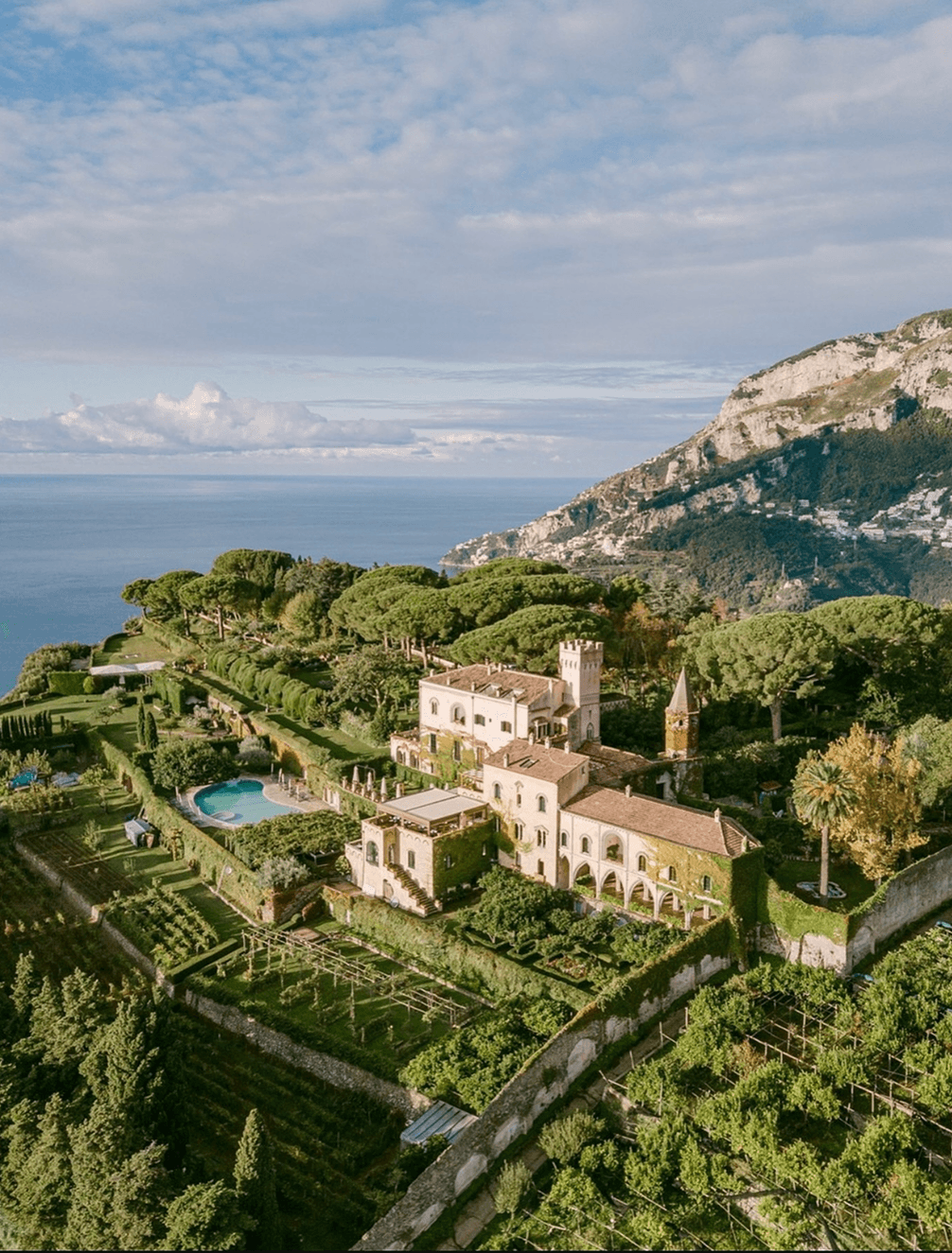Aerial view of a villa with a pool surrounded by lush greenery, overlooking the sea and a mountain landscape under a partly cloudy sky.