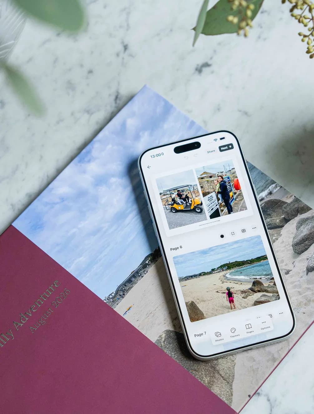 Smartphone displaying vacation photos from a trip, resting on a memory book labeled "Family Adventure August 2026" on a marble surface.