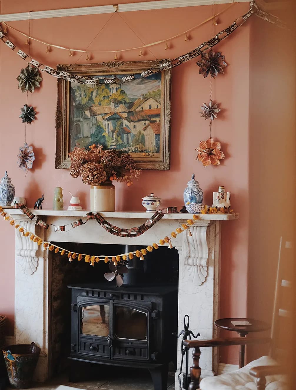 A cozy living room with a decorated marble fireplace, featuring colorful garlands, an autumn bouquet, ornaments, and a painting above.