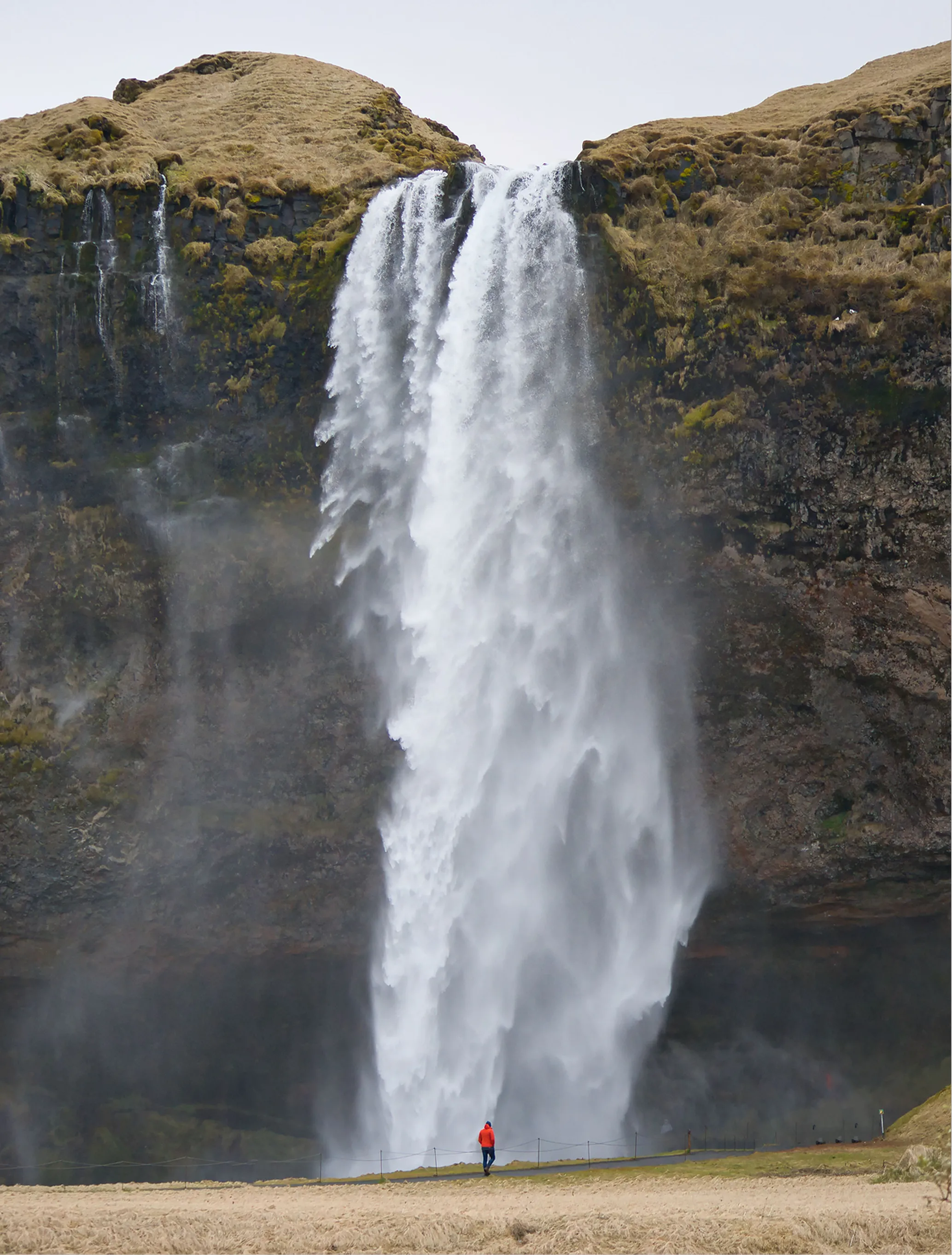 Seljalandsfoss waterfall in Iceland cascades down a cliff face, with a person in an orange jacket walking near its misty base.