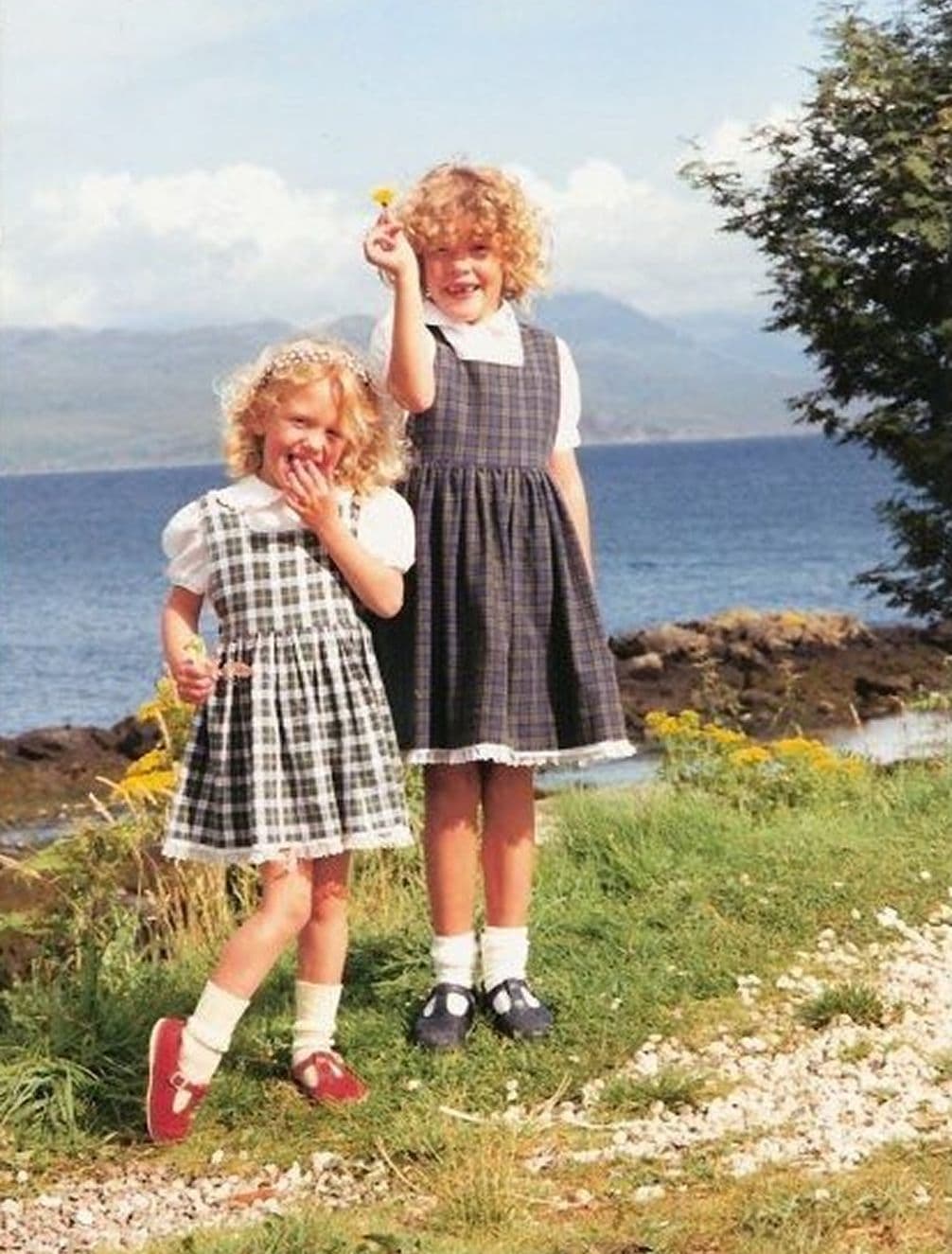 Two young girls with curly hair wearing plaid dresses stand on a grassy area by a scenic lakeside, one holding a yellow flower.