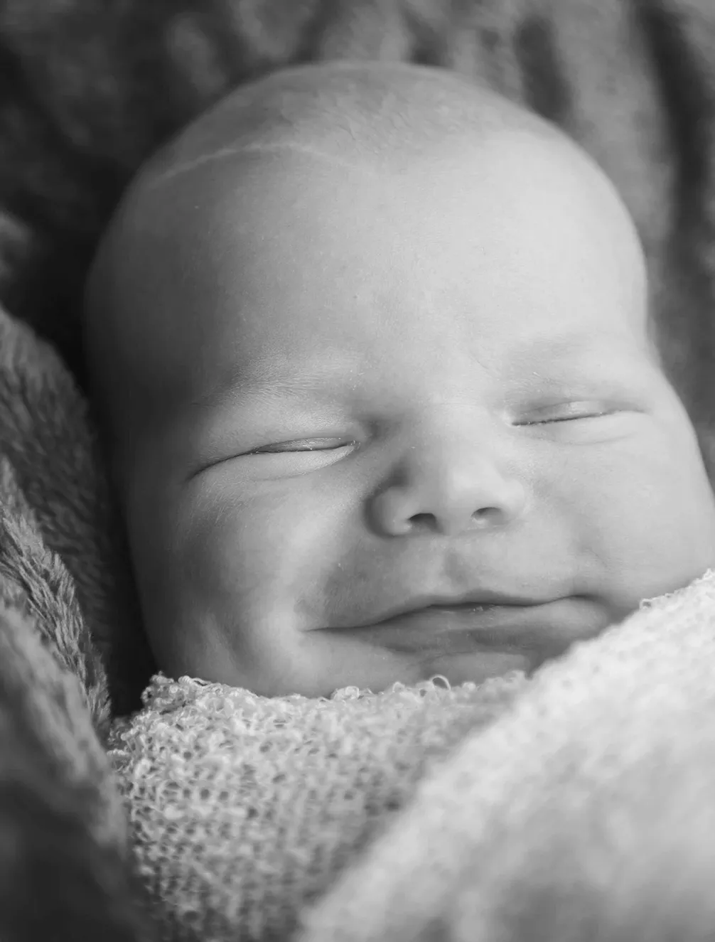 Close-up of a smiling newborn baby wrapped in a knitted blanket, eyes gently closed, in black and white.