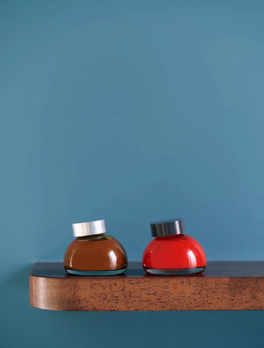 Two small, round glass bottles, one brown with a silver cap and one red with a black cap, sit on a wooden shelf against a blue wall.