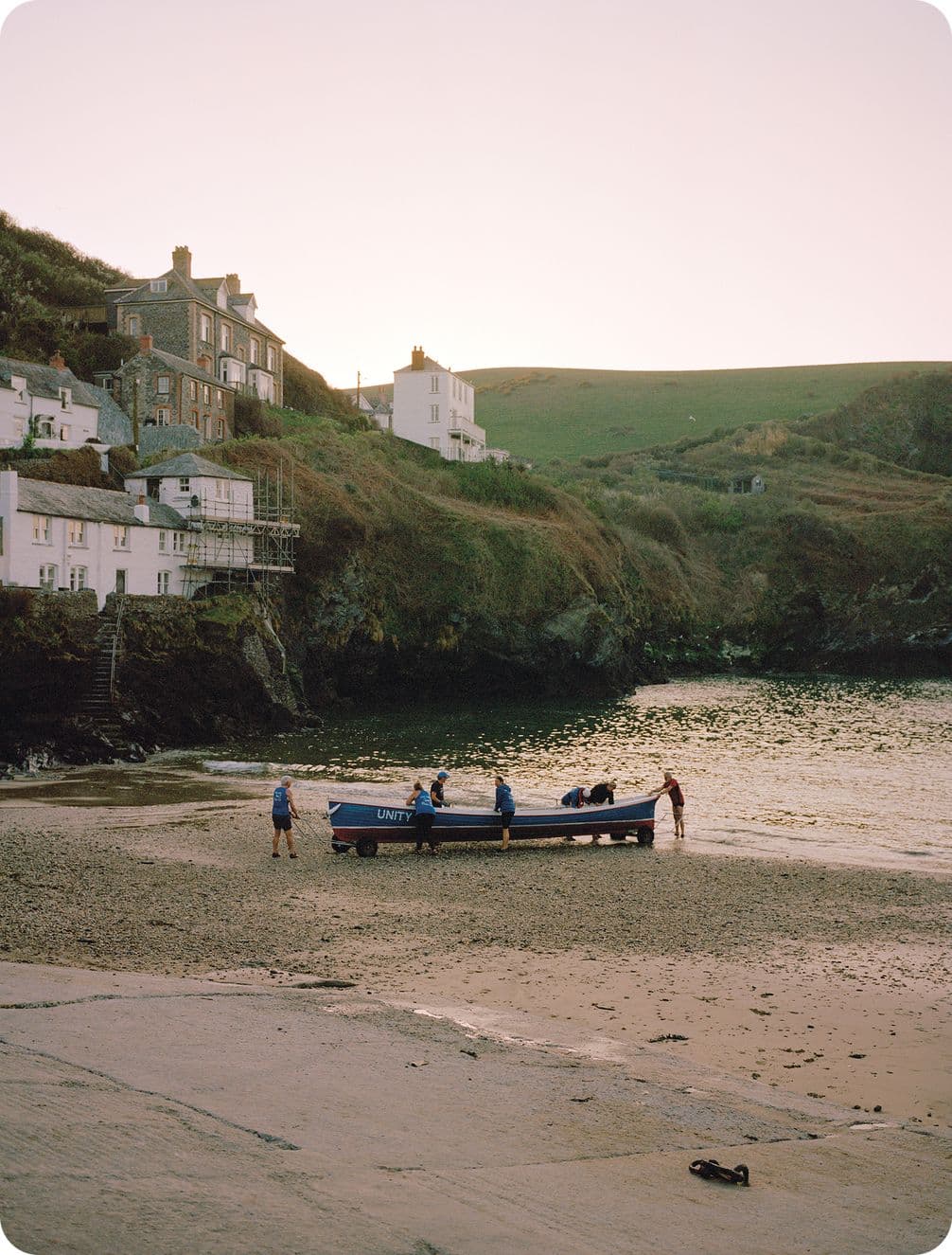 A small boat with people is on a sandy beach near calm water, surrounded by cliffs and houses under a soft, pinkish sky.