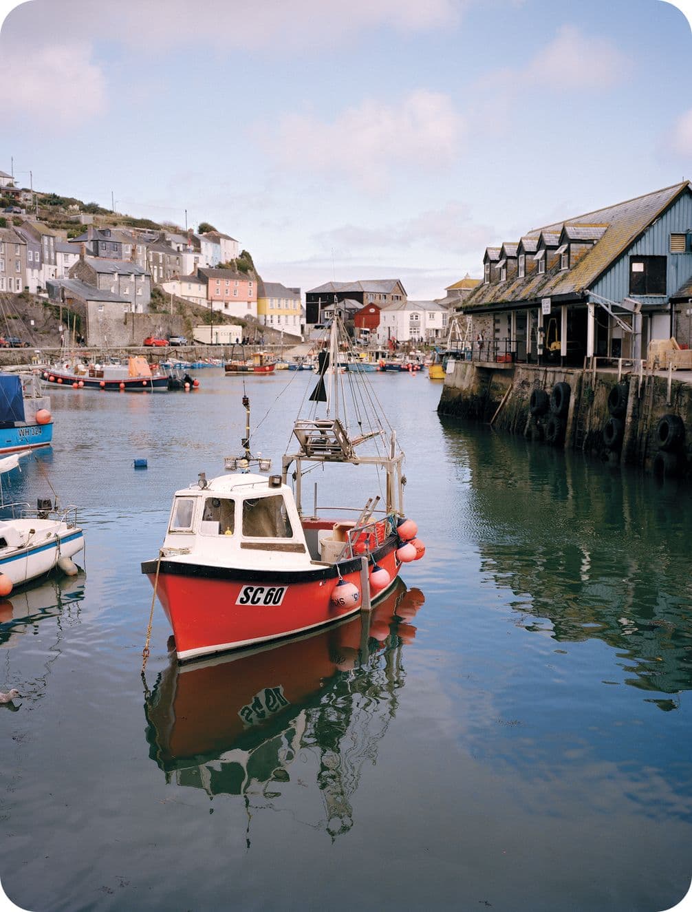 A small red fishing boat floats in a calm harbor, surrounded by quaint buildings and a clear sky.