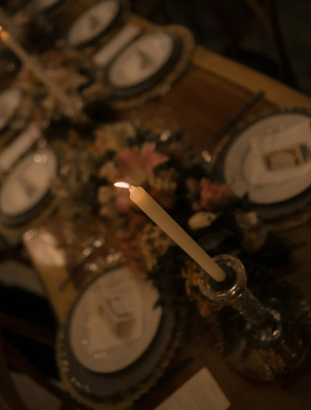 A dimly lit dinner table with a lit candle, floral arrangement, and intricately set plates, creating an elegant and intimate atmosphere.