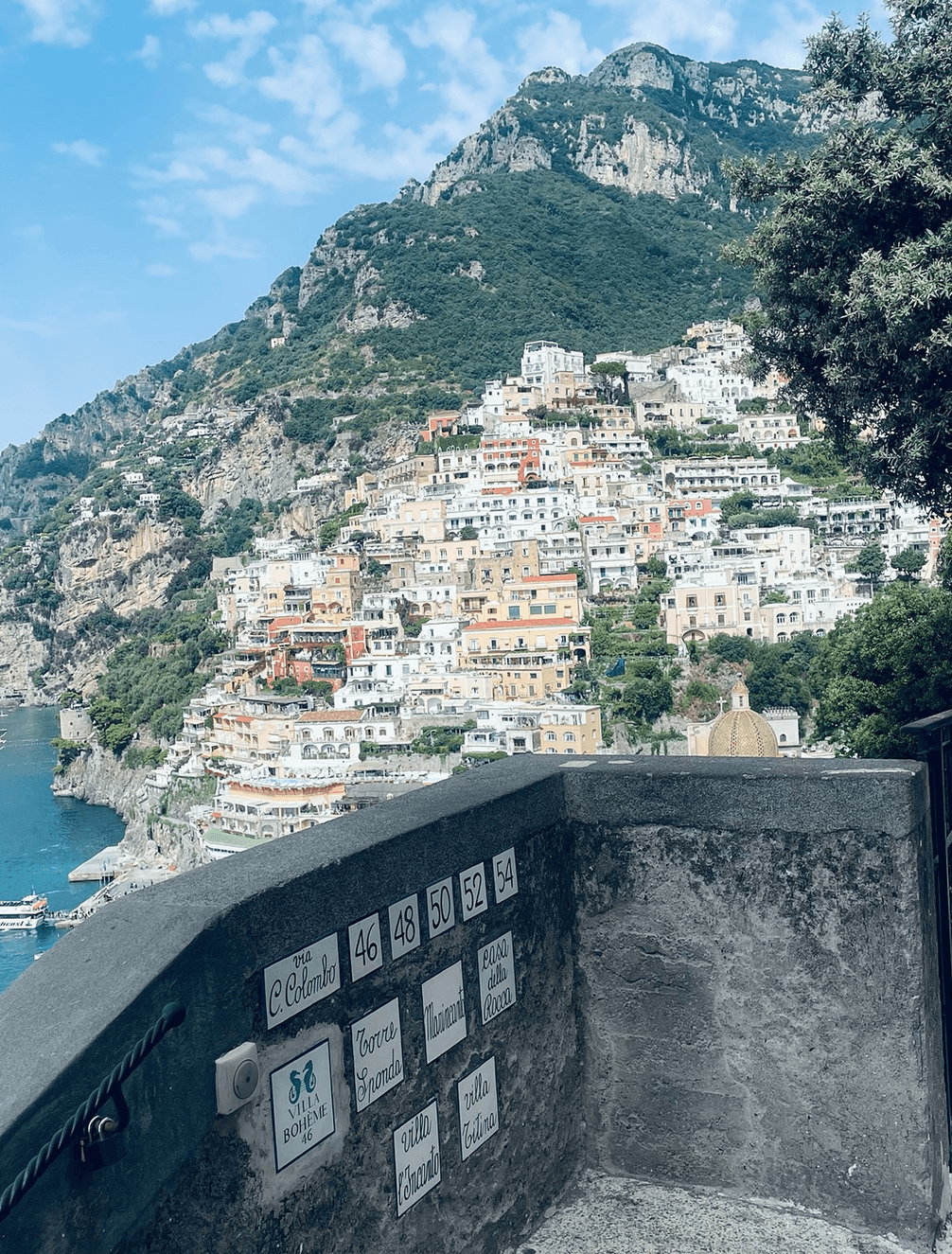 Colorful cliffside buildings overlooking the sea in Positano, Italy, with a stone wall and directional signs in the foreground.