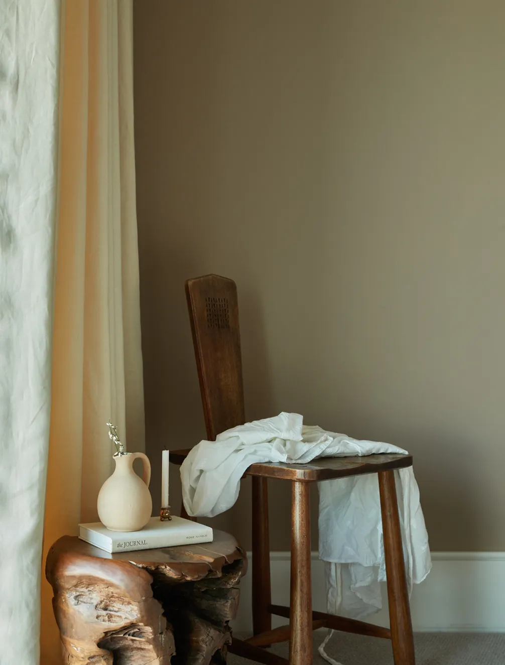 A wooden chair with a white fabric draped over it, next to a wooden stool with stacked books and a small vase, near beige curtains.