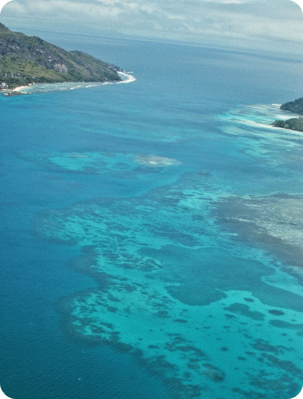Aerial view of turquoise ocean with coral reefs and sandy shallows, flanked by green coastal hills under a cloudy sky.