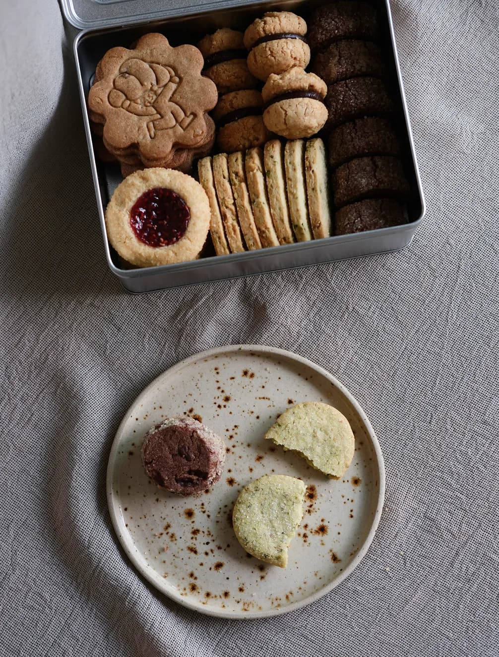 An open tin of assorted cookies with a round plate below; two cookies, one chocolate and one partially eaten.