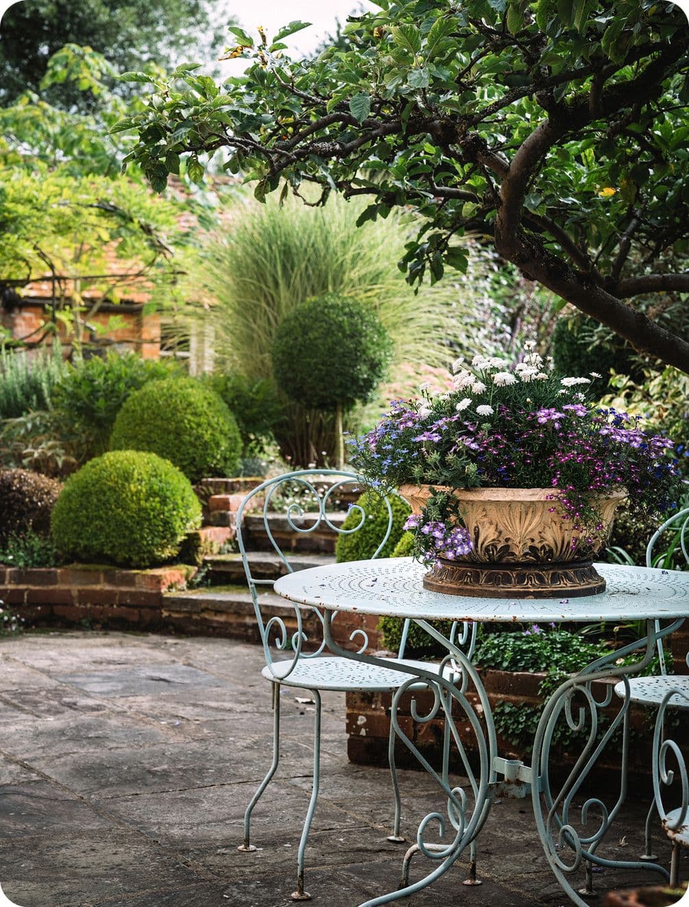 A quaint garden with a white metal table and chairs, adorned with a floral centerpiece, surrounded by lush greenery and trimmed bushes.