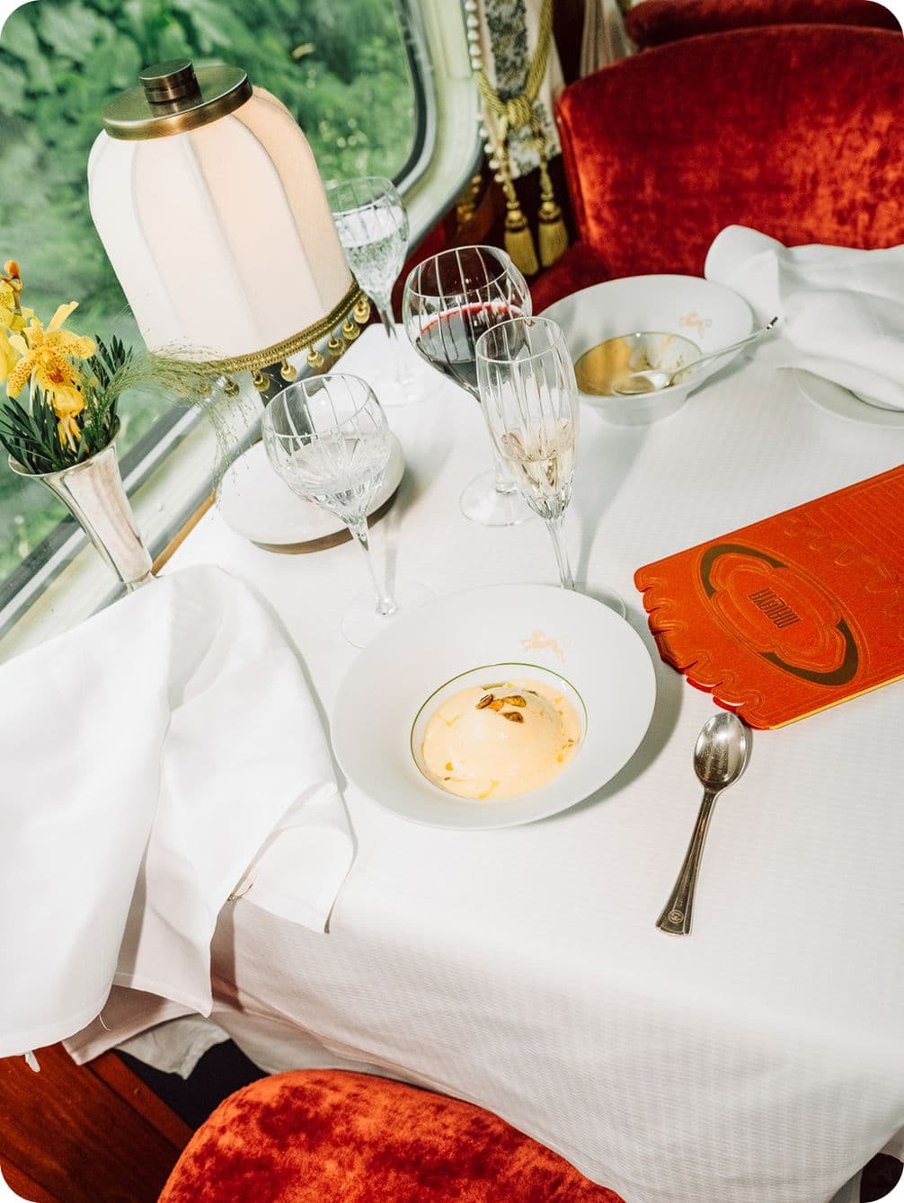 Elegant dining setup on a train with white tablecloth, crystal glasses, a lamp, and a dessert bowl, surrounded by red velvet seats.